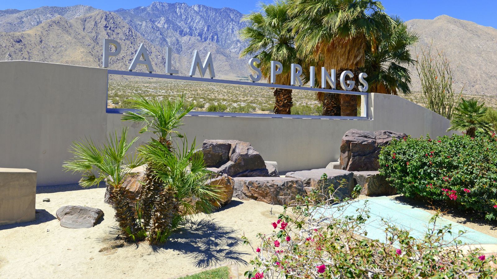 A "Palm Springs" sign stands amid desert plants and palm trees, with mountains in the background.