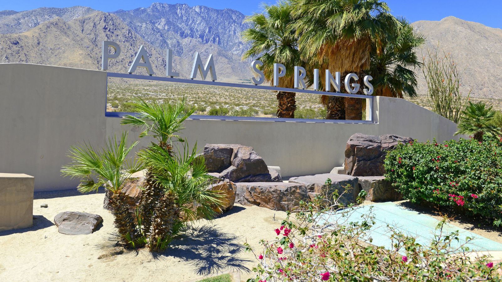 A "Palm Springs" sign stands before desert landscaping, palm trees, rocks, and mountains under a clear blue sky.