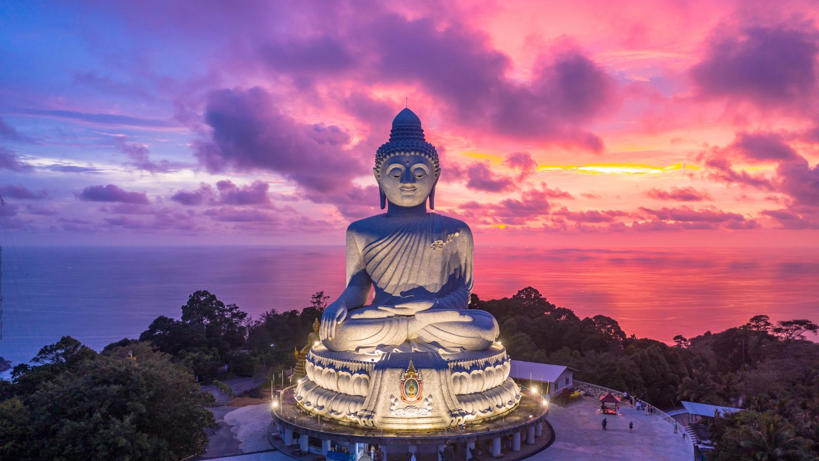 Large Buddha statue sits on a hilltop above trees, with a pink-purple sunset sky reflecting over the ocean behind it.