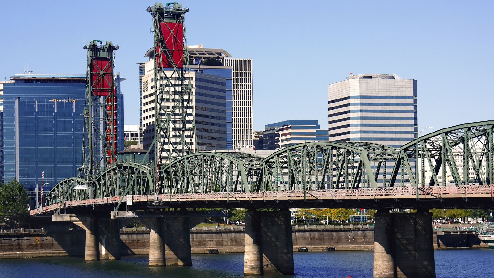 A steel truss bridge crosses a river before modern office buildings under a clear sky.
