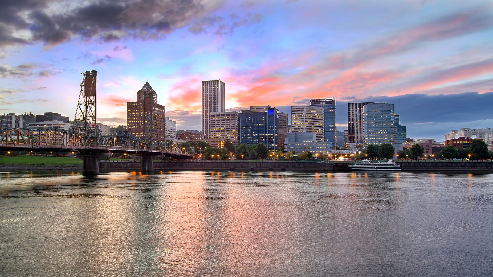 City skyline with tall buildings and a bridge over a river at sunset, colorful clouds reflected in the water.