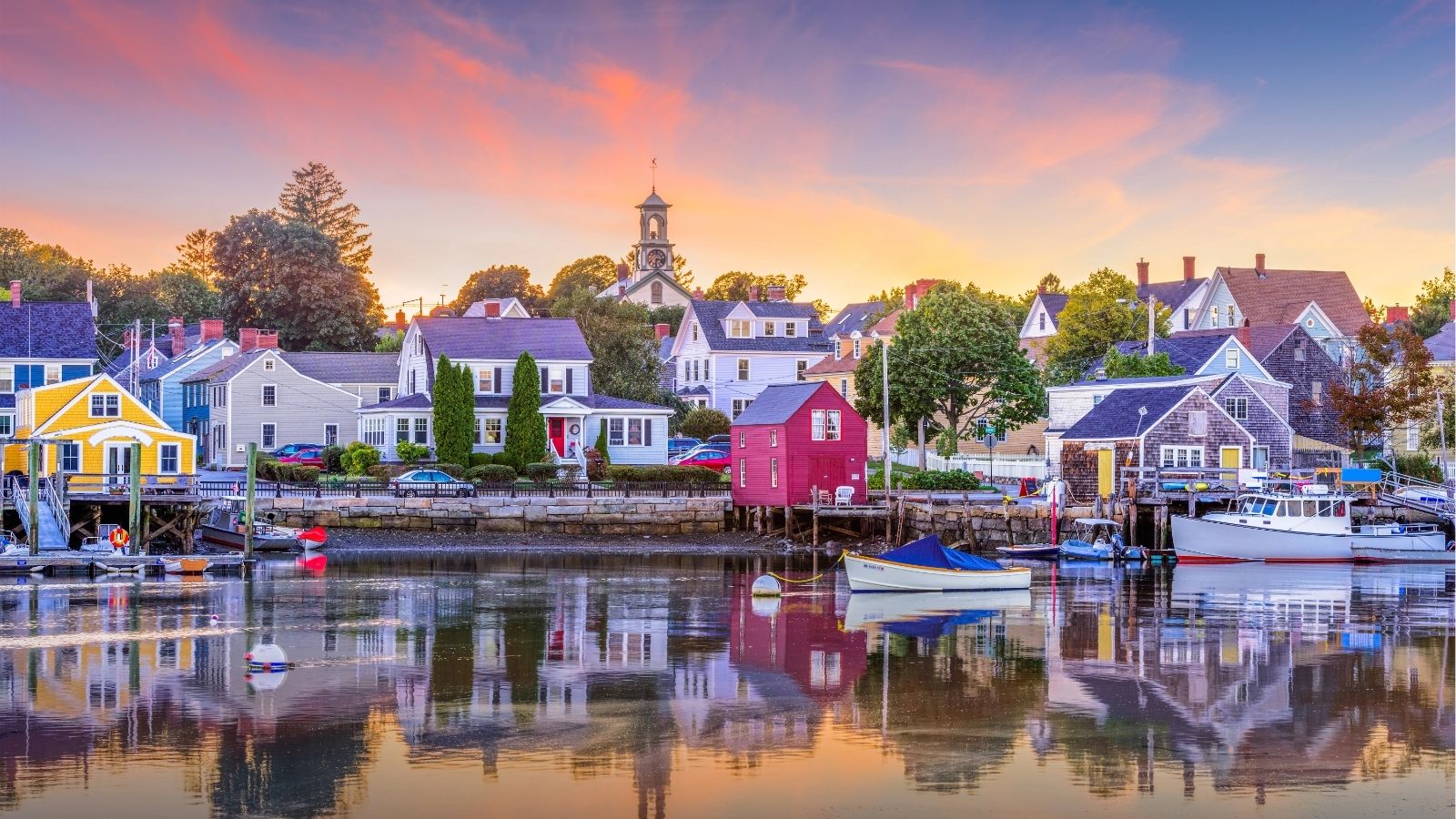 Sunset harbor with colorful houses, docked boats, and a church steeple in the background.