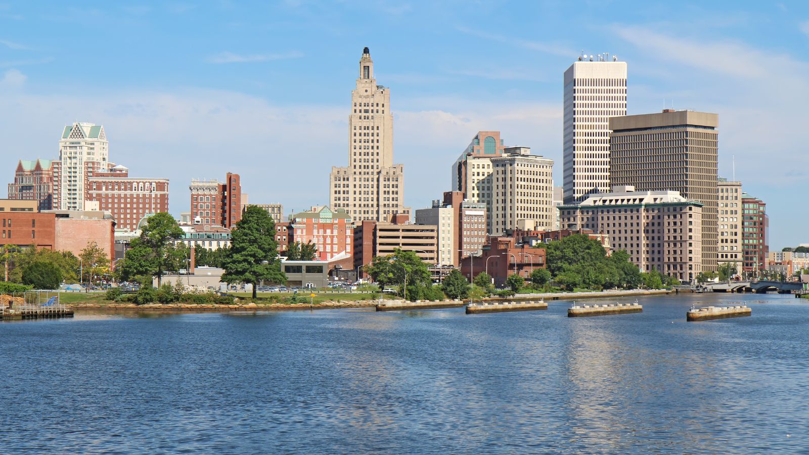 City skyline with tall buildings, trees, and water beneath a partly cloudy blue sky.