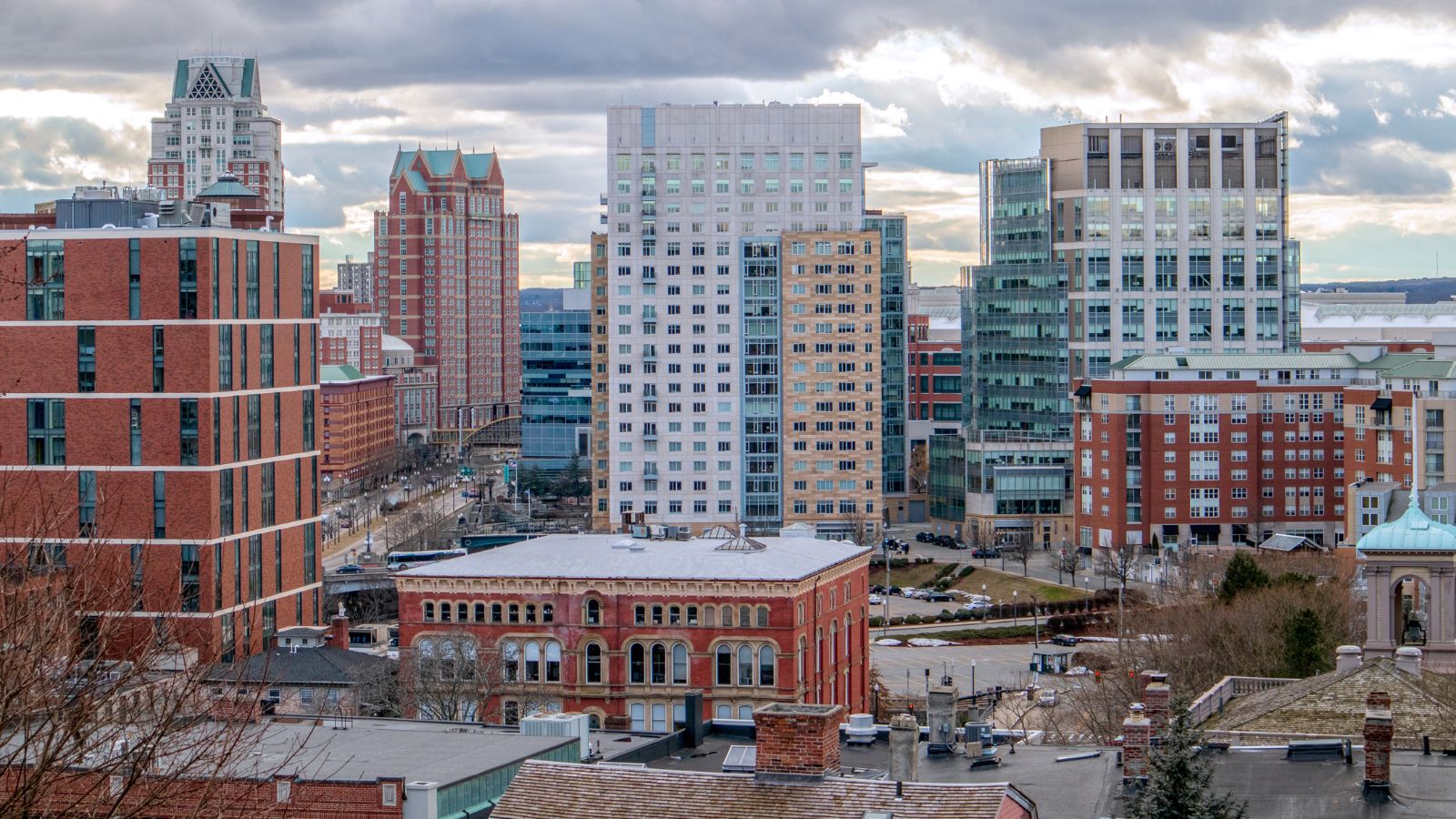 A cityscape with mid- and high-rise brick and glass buildings beneath a cloudy sky.