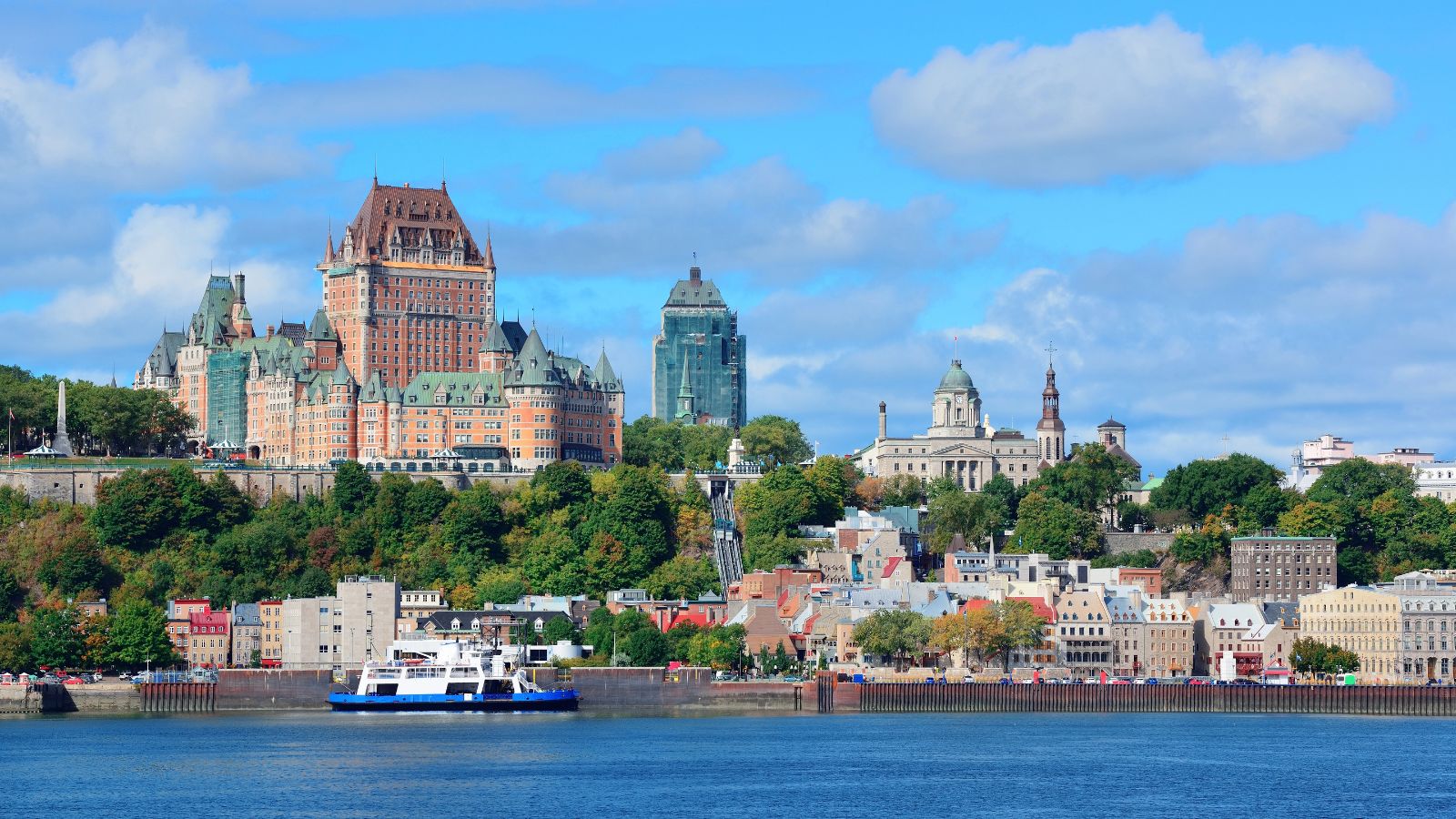 A ferry crosses the river before Quebec City's skyline, with Château Frontenac, historic buildings, and greenery below a cloudy sky.