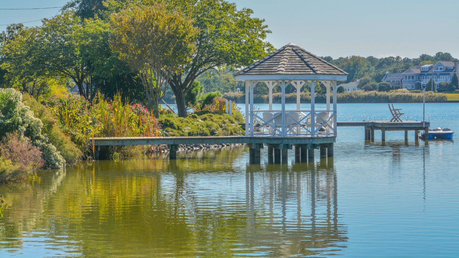 A white gazebo stands on a wooden dock over a calm lake, surrounded by greenery, with houses and another dock in the background.