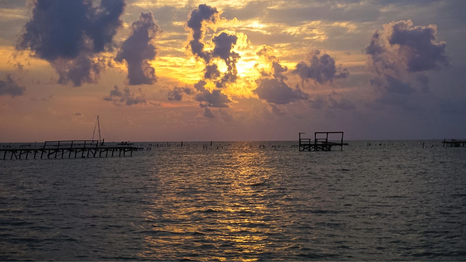 Sunset over calm water with wooden piers, a distant sailboat, and clouds reflecting golden light on the surface.