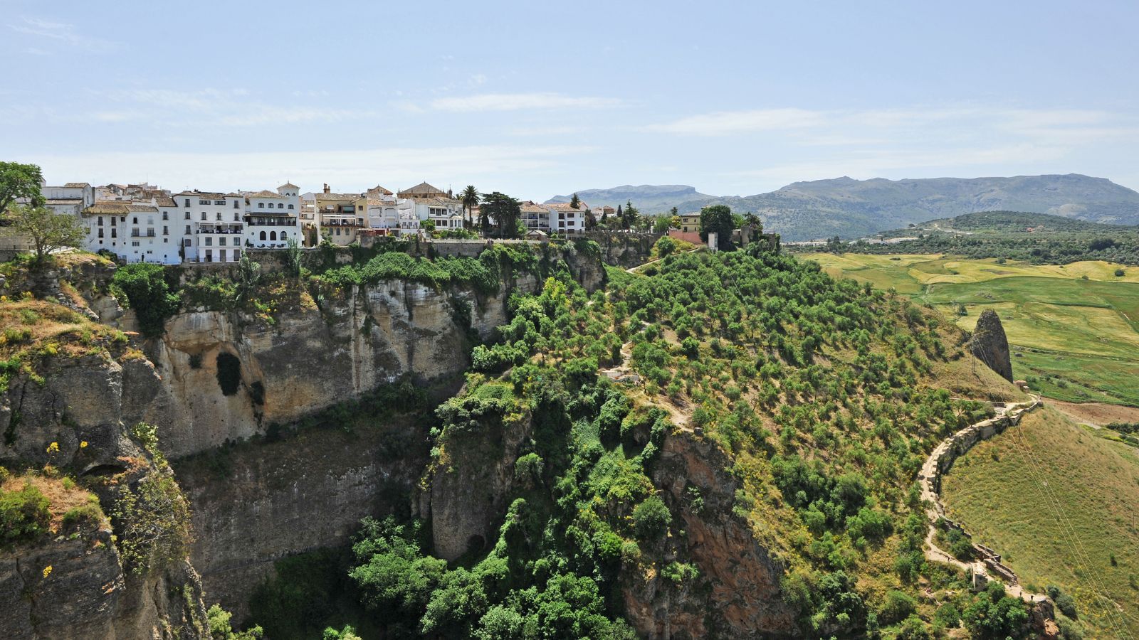 White buildings perch on cliffs above a green valley and winding path, with mountains in the background under a clear sky.