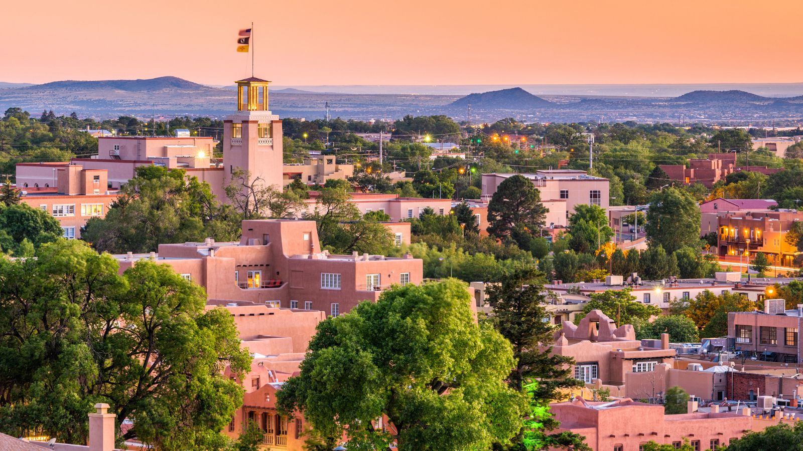 Santa Fe, New Mexico at sunset with adobe buildings, green trees, and distant mountains beneath an orange sky.