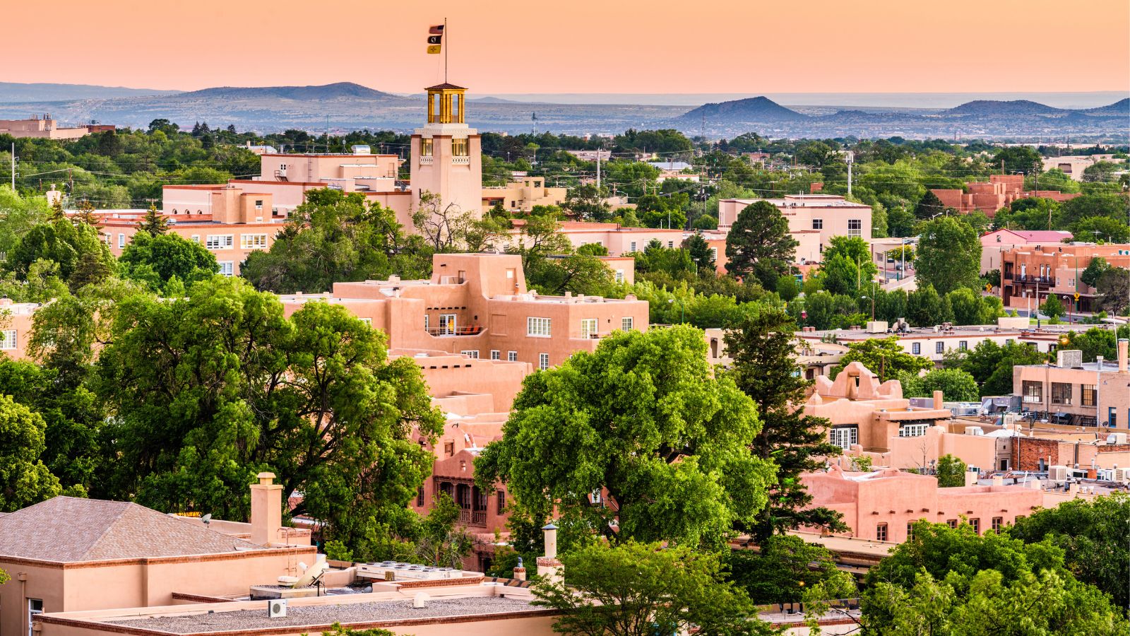 Adobe-style buildings, green trees, a clock tower with a flag, and distant hills under an orange sky in Santa Fe, New Mexico.