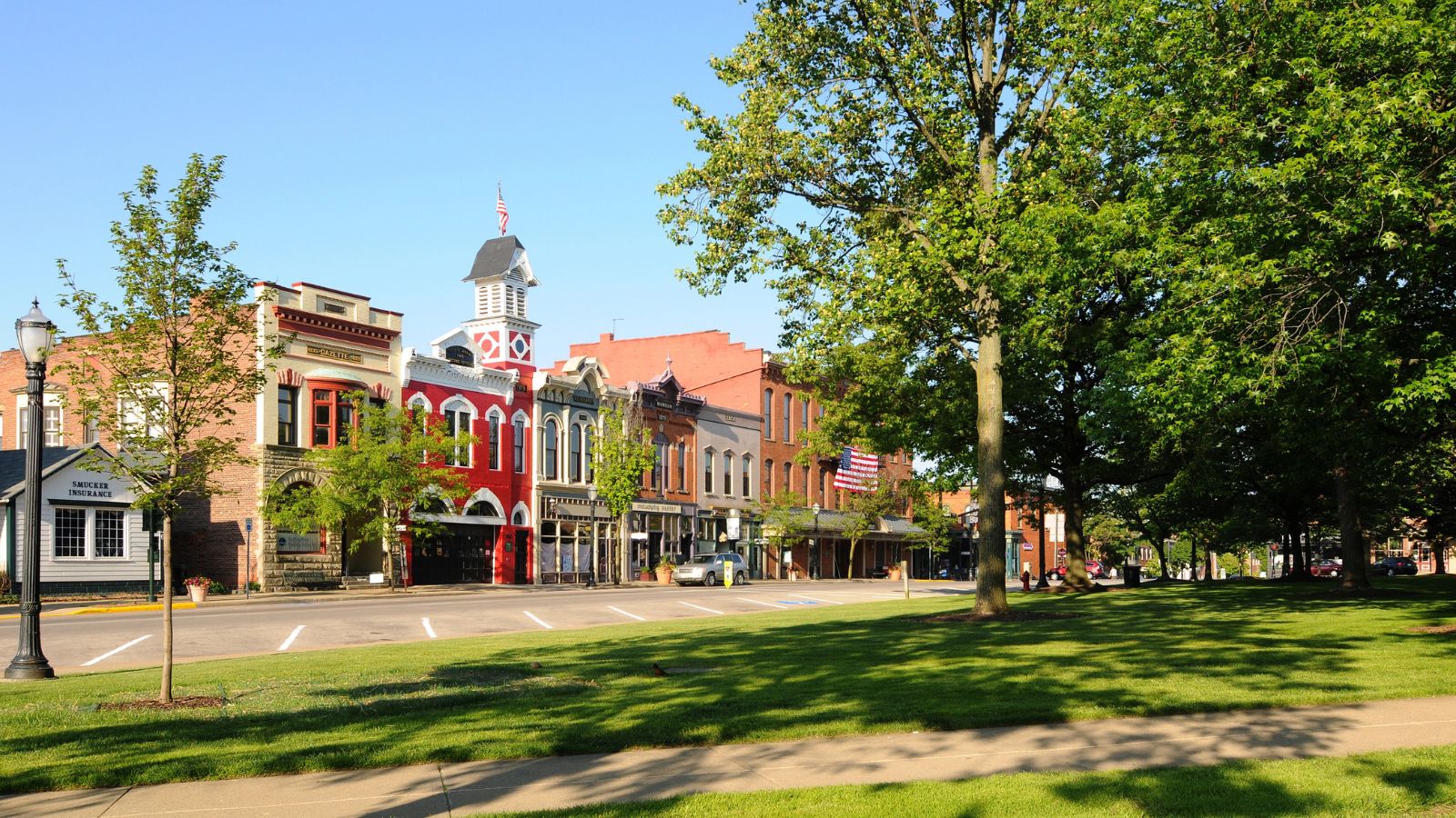 Historic brick buildings line a quiet small-town street, with green trees and a grassy park beneath a clear blue sky.