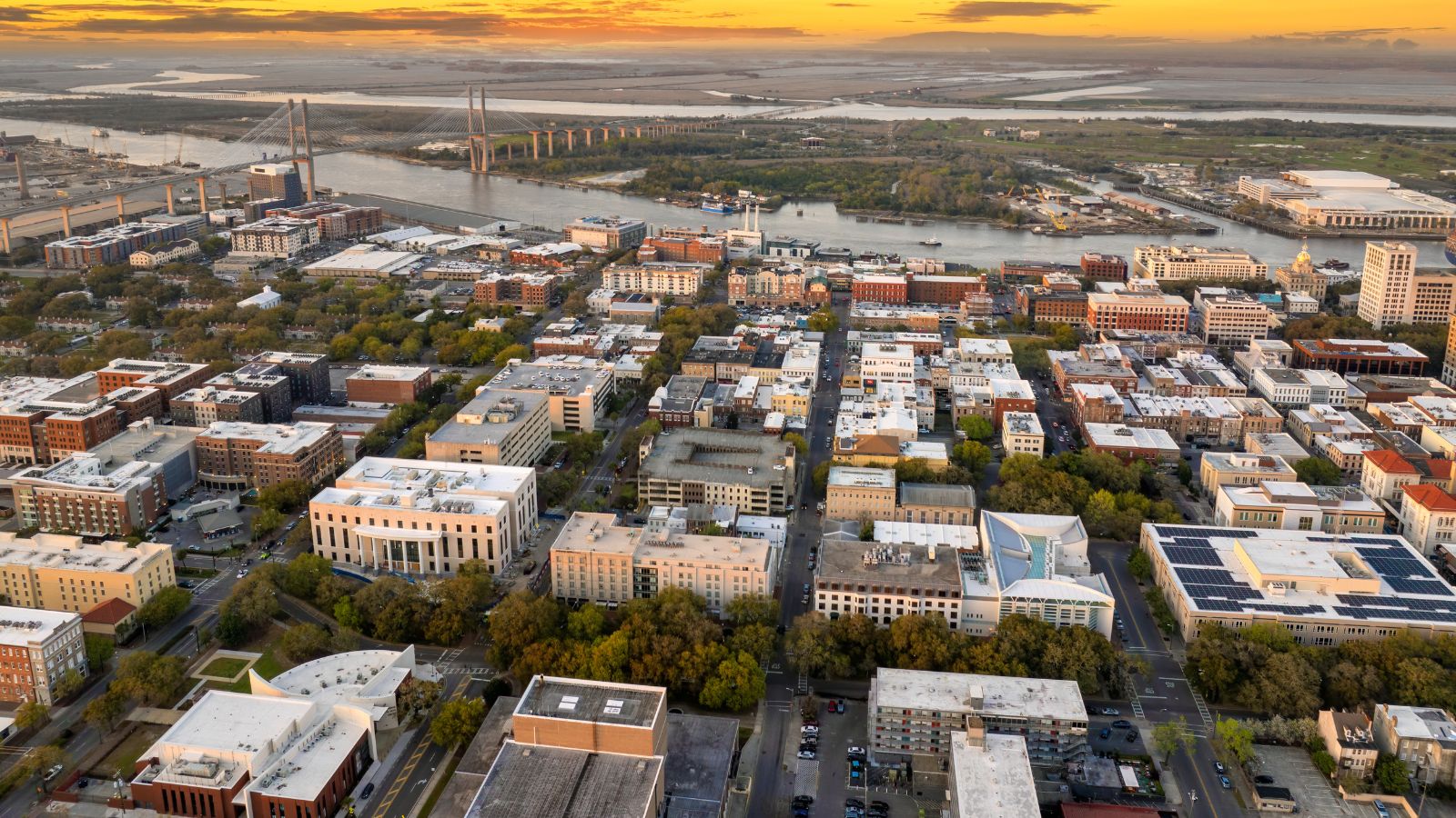 Aerial city view at sunset with a river, bridge, grid-patterned streets, buildings, and green spaces.