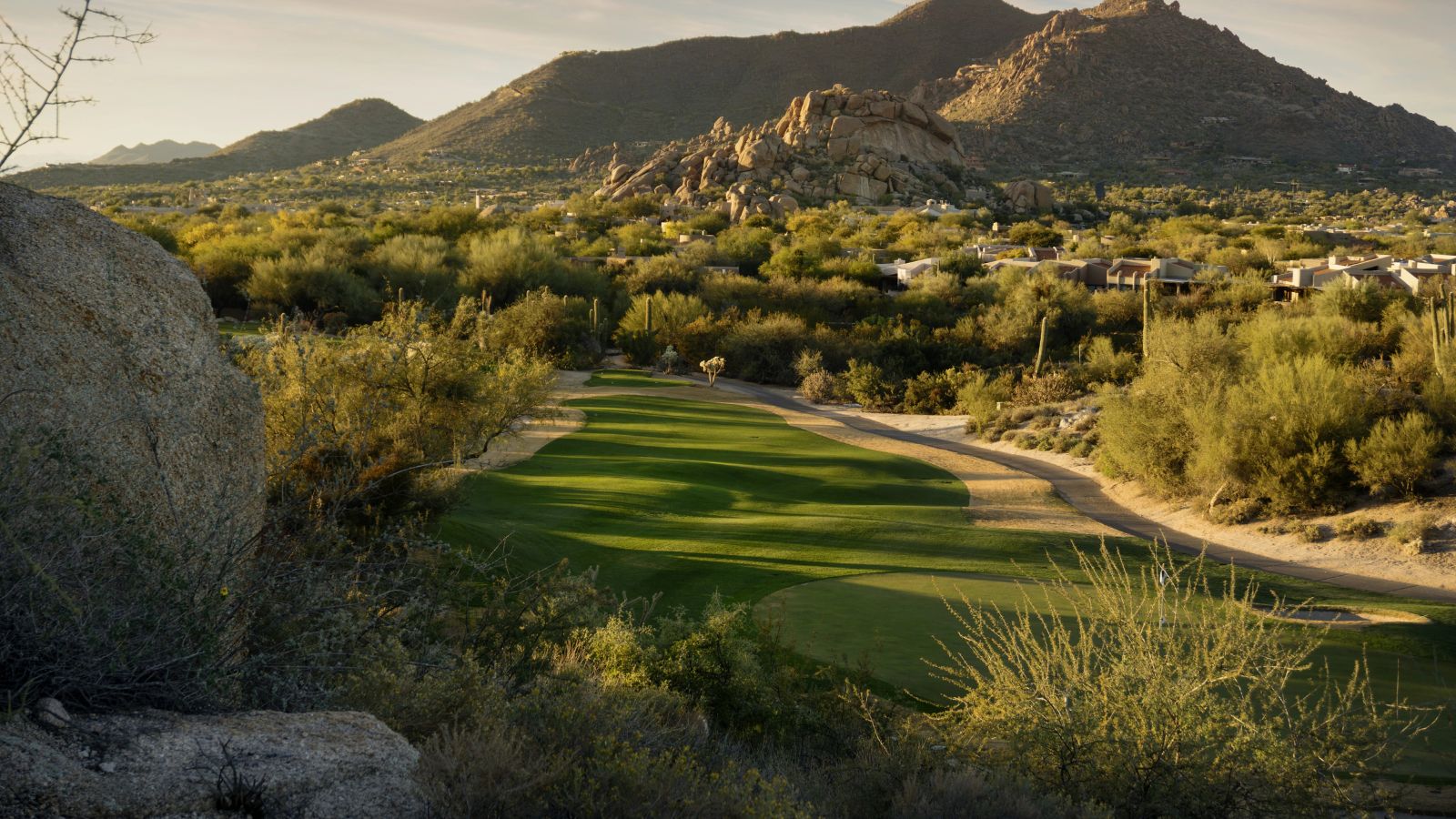 A golf course fairway winds through desert shrubs and rocks, with mountains and a partly cloudy sky in the background.