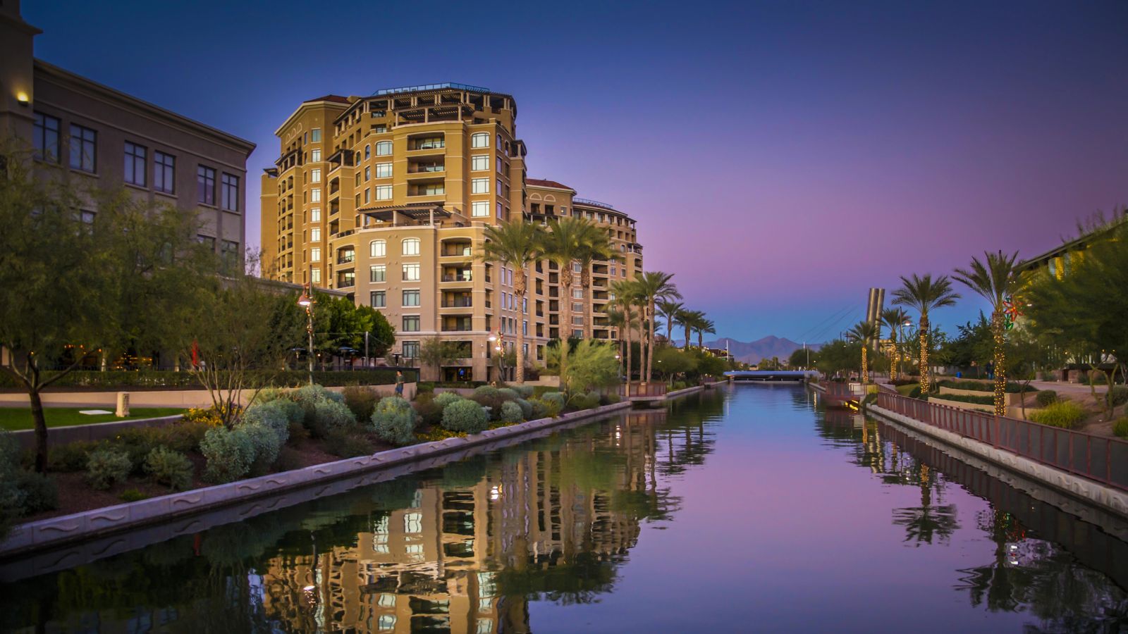 At dusk, a multistory building and palm trees are reflected in a canal beneath a purple-blue sky.