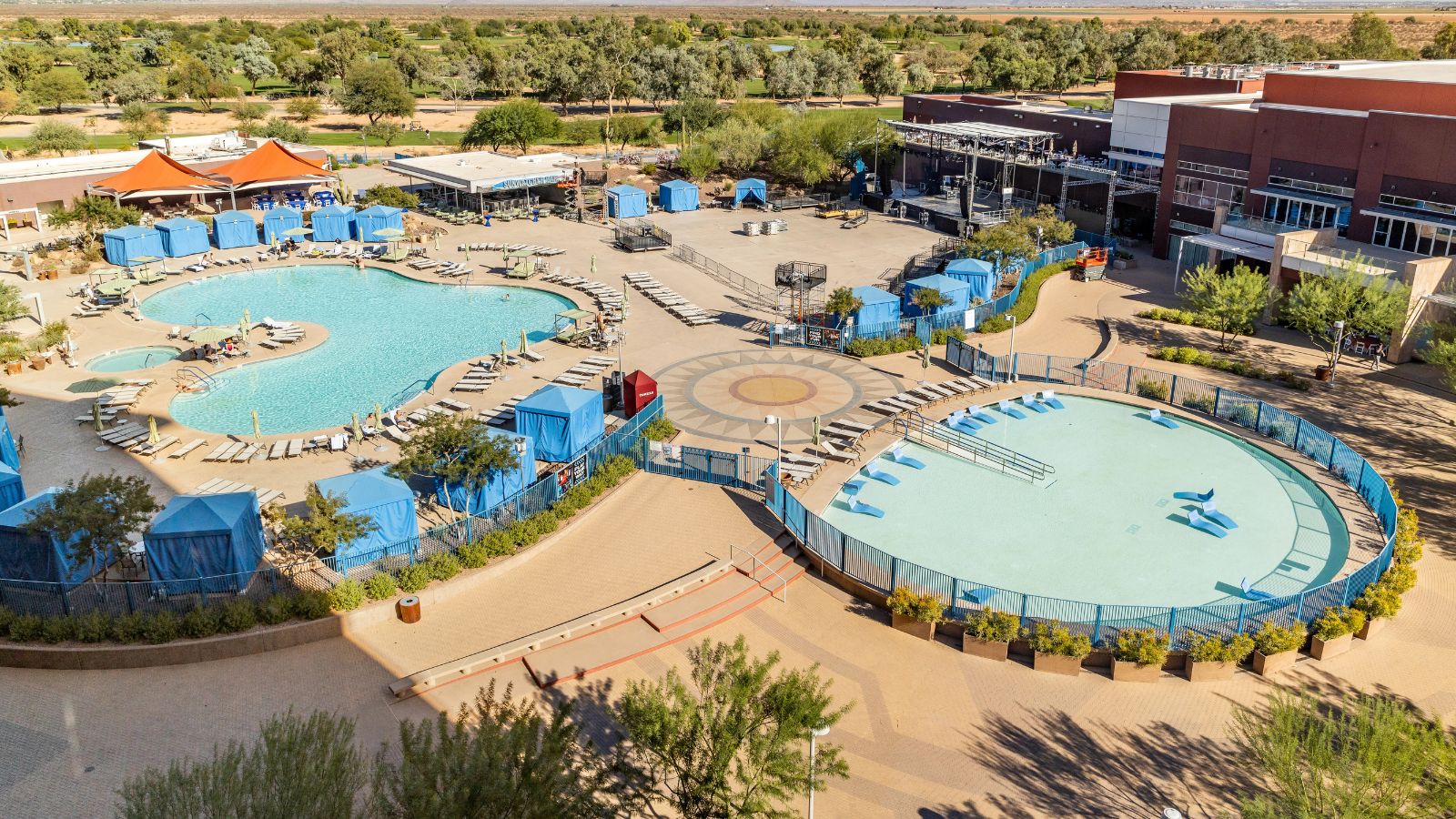 Aerial view of a resort with two pools, blue cabanas, lounge chairs, adjacent buildings, and surrounding trees.