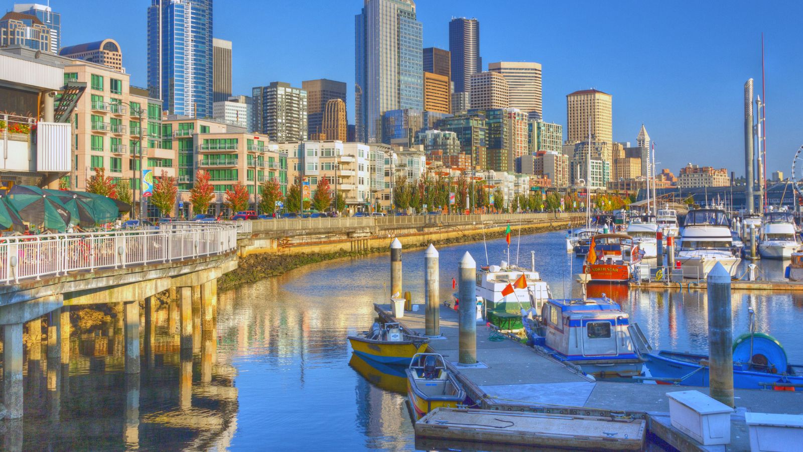 Boats are docked at a marina with modern skyscrapers in the background beneath a clear blue sky.