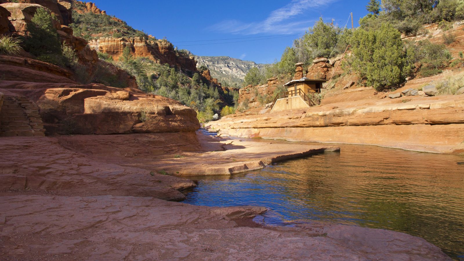 A clear stream winds through a red rock canyon with sparse plants and a stone building on the right beneath a bright blue sky.