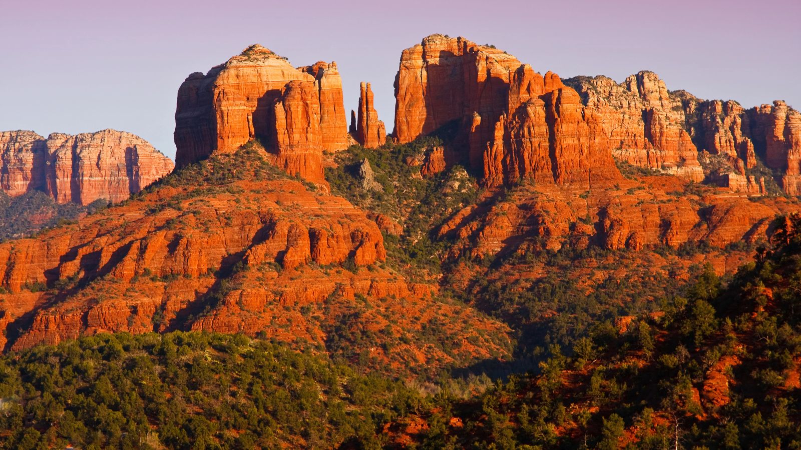 Red rock formations tower over green vegetation beneath a clear sky, all illuminated by warm sunlight.
