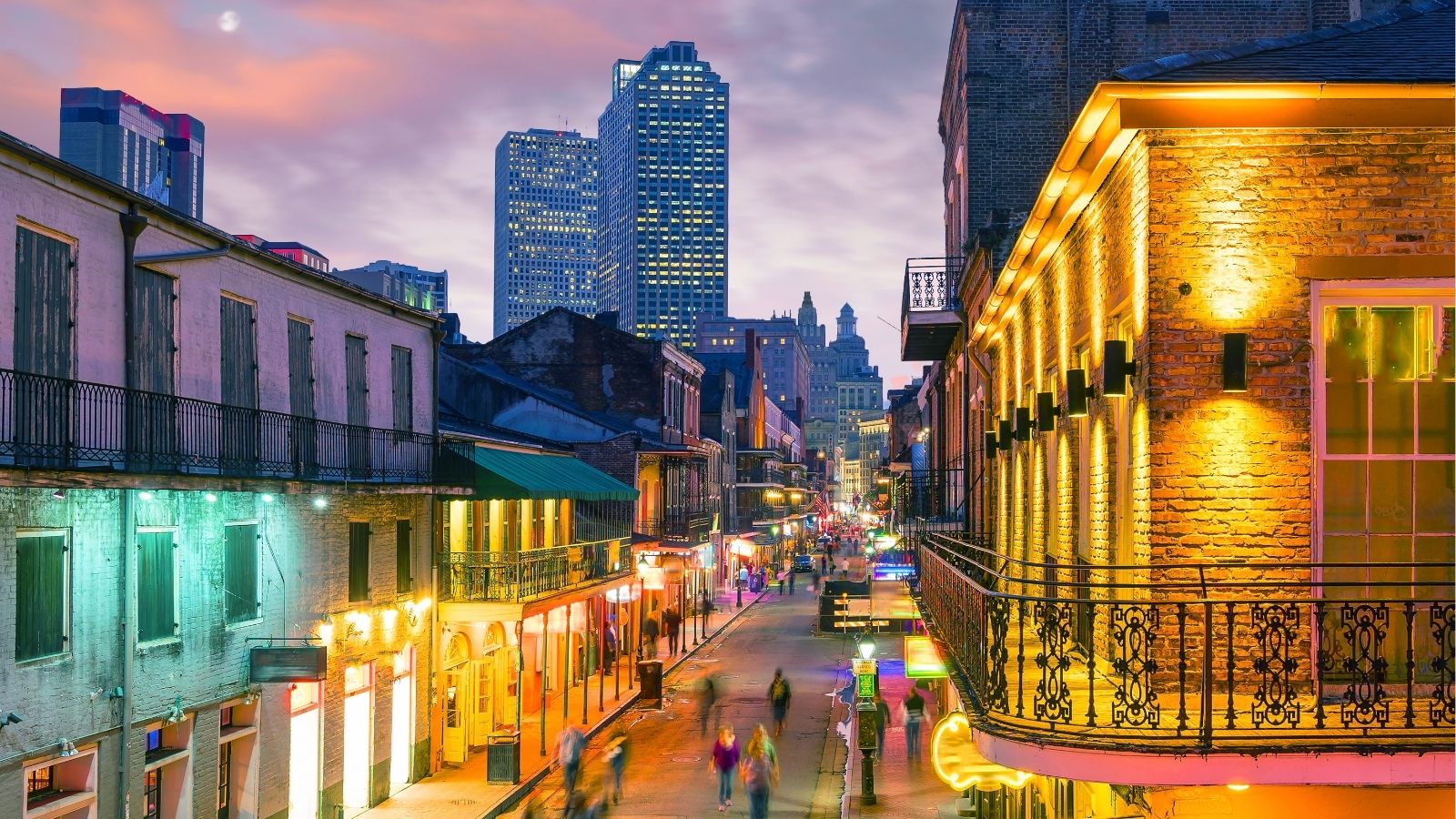 City street at dusk with blurred pedestrians, historic brick buildings, and skyscrapers beneath a colorful evening sky.