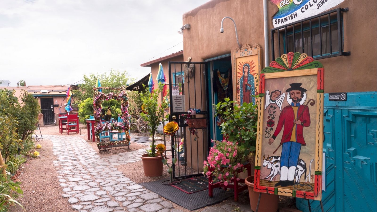 Cobblestone walkway passes folk art, flowers, and a painted sign outside a Southwestern adobe-style shop.