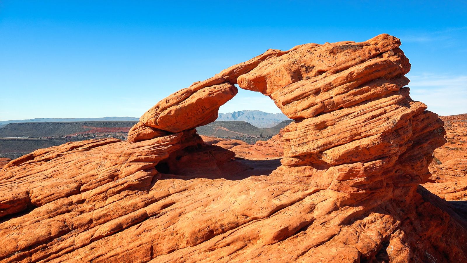 A red sandstone arch rises beneath a blue sky, framing distant mountains through its central opening.