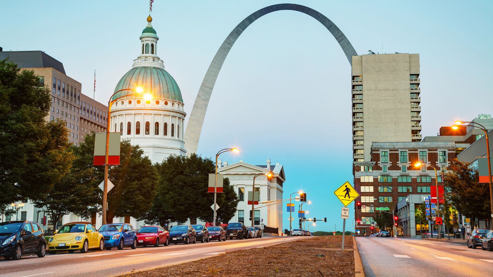 Downtown St. Louis at dusk with the Gateway Arch, Old Courthouse, and cars parked along the street.