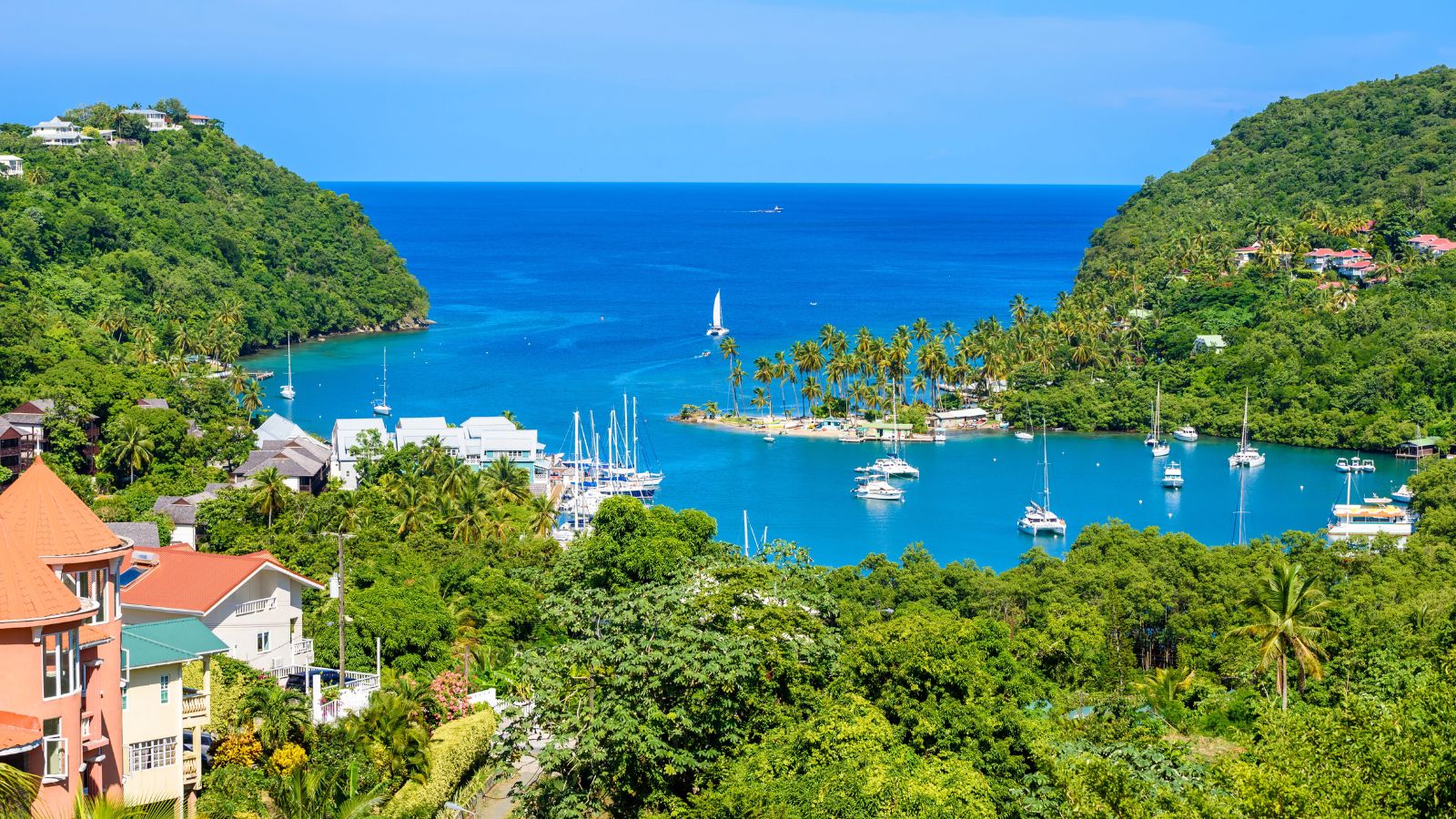 Aerial view of a tropical bay with anchored boats, green hills, and seaside buildings under a clear blue sky.