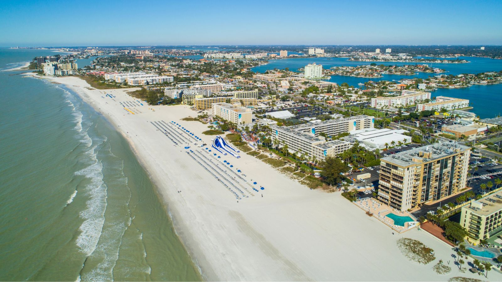 Aerial view of a sandy beach with beachfront hotels, waves, and a city with waterways under clear blue skies.