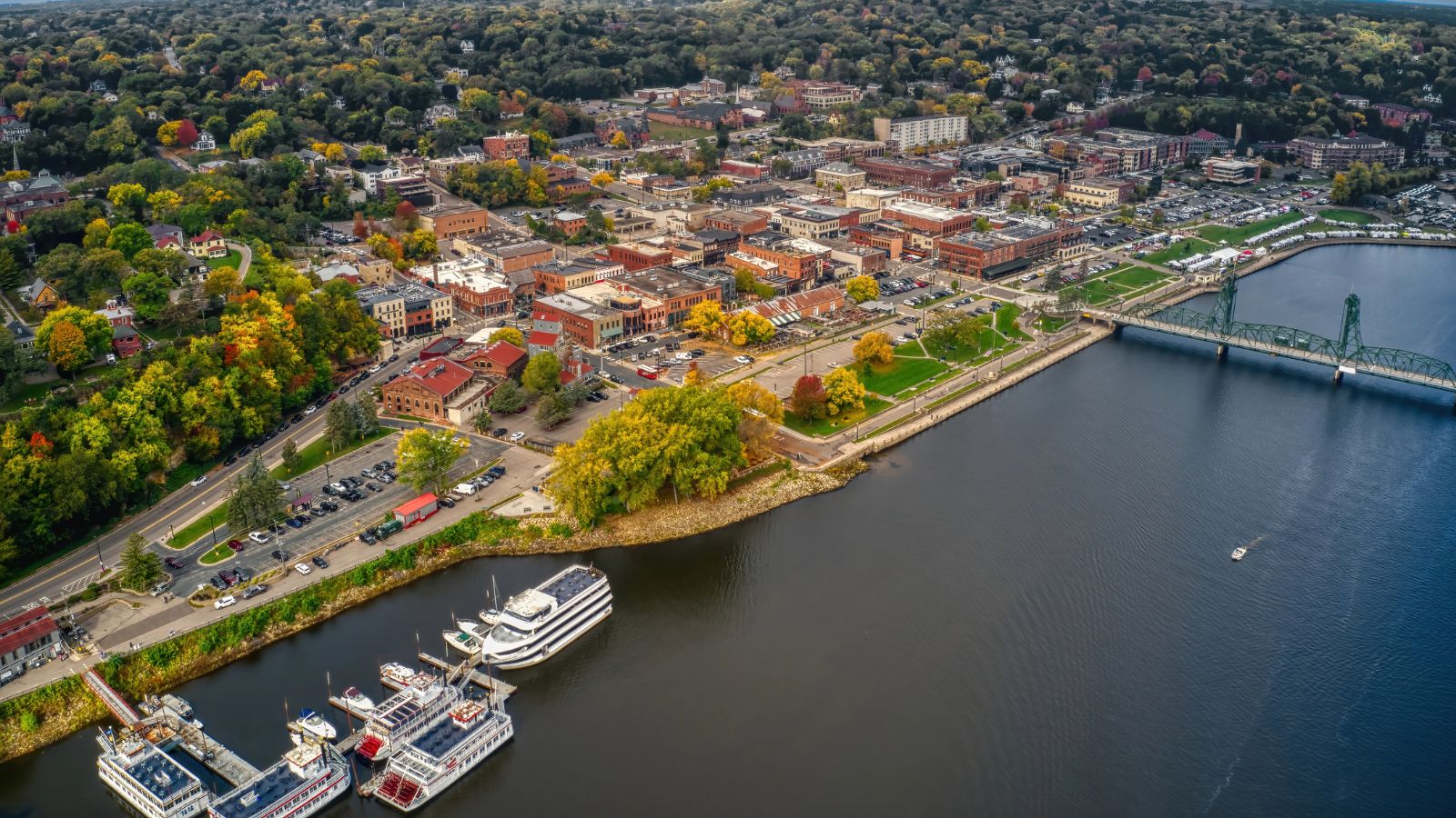 Aerial view of a small city with brick buildings, riverside docks, boats, a bridge, and autumn tree-lined streets.