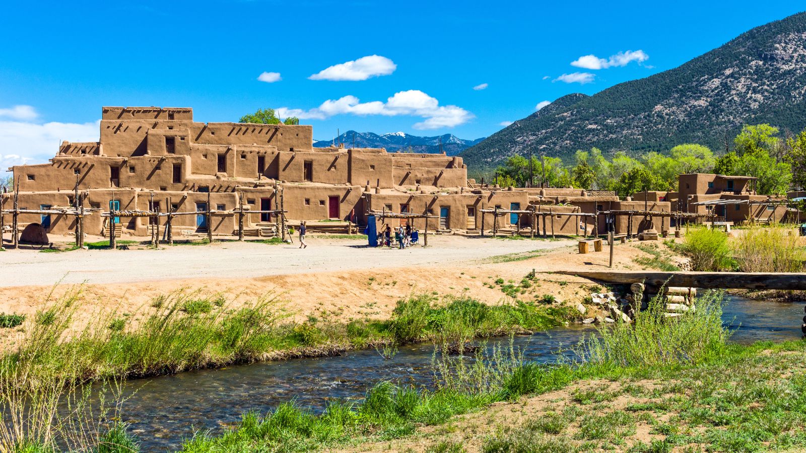 Taos Pueblo’s multi-story adobe buildings stand by a stream at the mountain’s base under a clear blue sky.