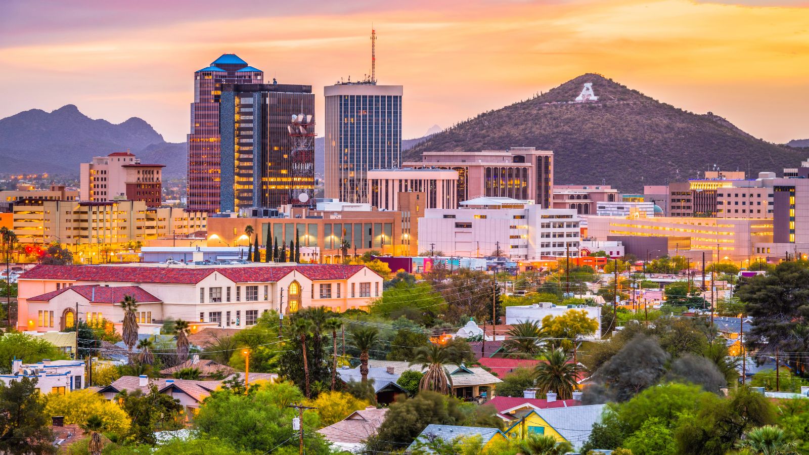 Downtown skyline with modern buildings, a church, and mountains at sunset; a large white "A" appears on the hillside.