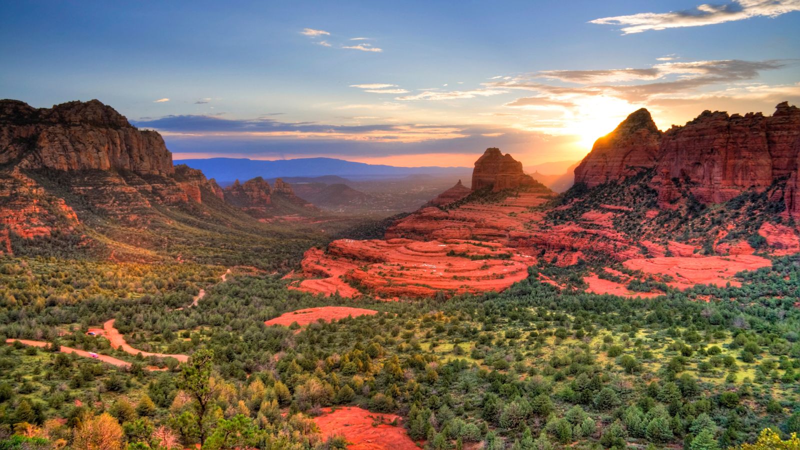 Sunset casts light on red rock formations and green vegetation in a desert beneath a partly cloudy sky.
