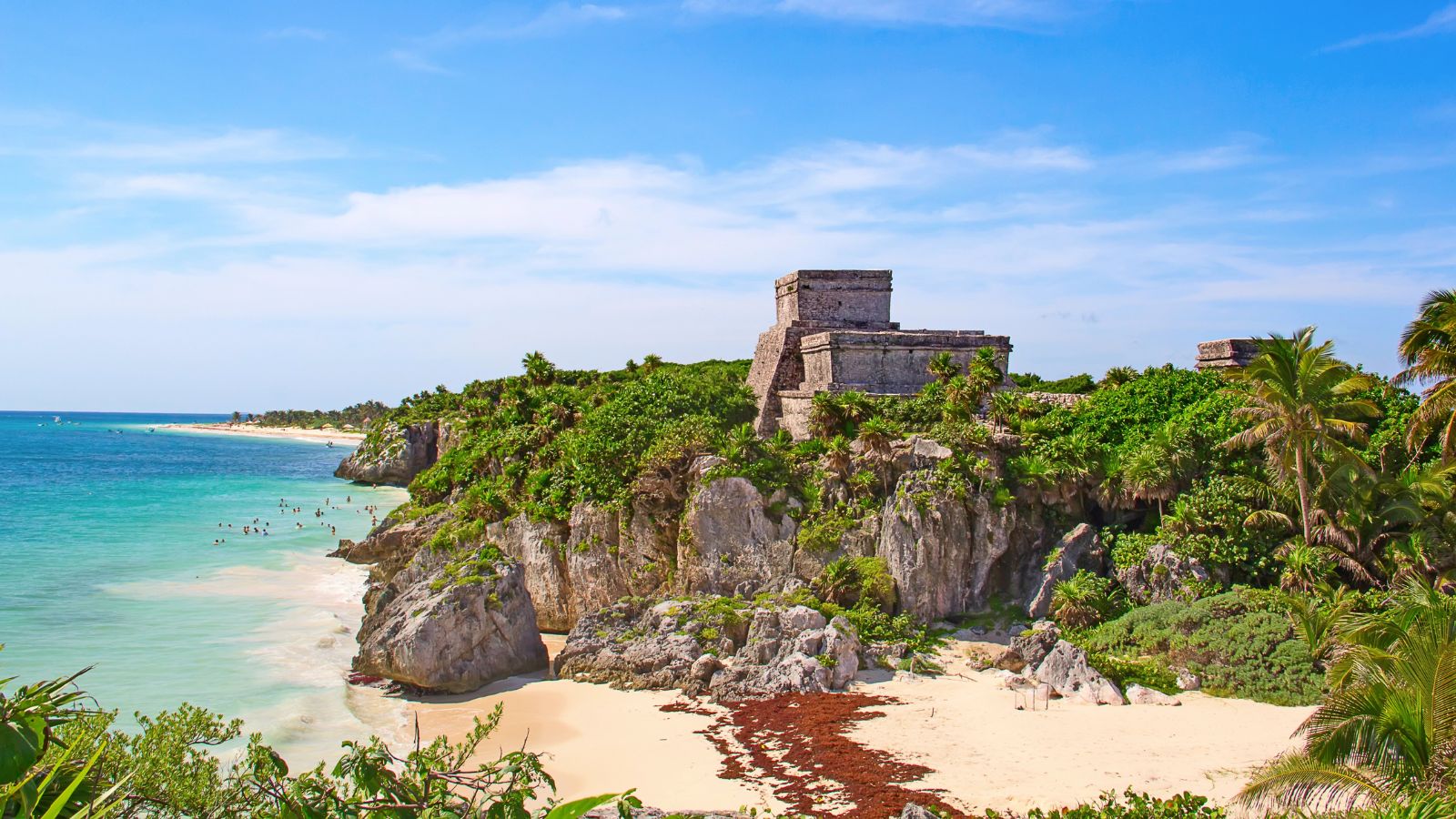 Ancient stone ruins rest on a rocky cliff above a sandy beach and turquoise ocean, bordered by lush greenery under a blue sky.