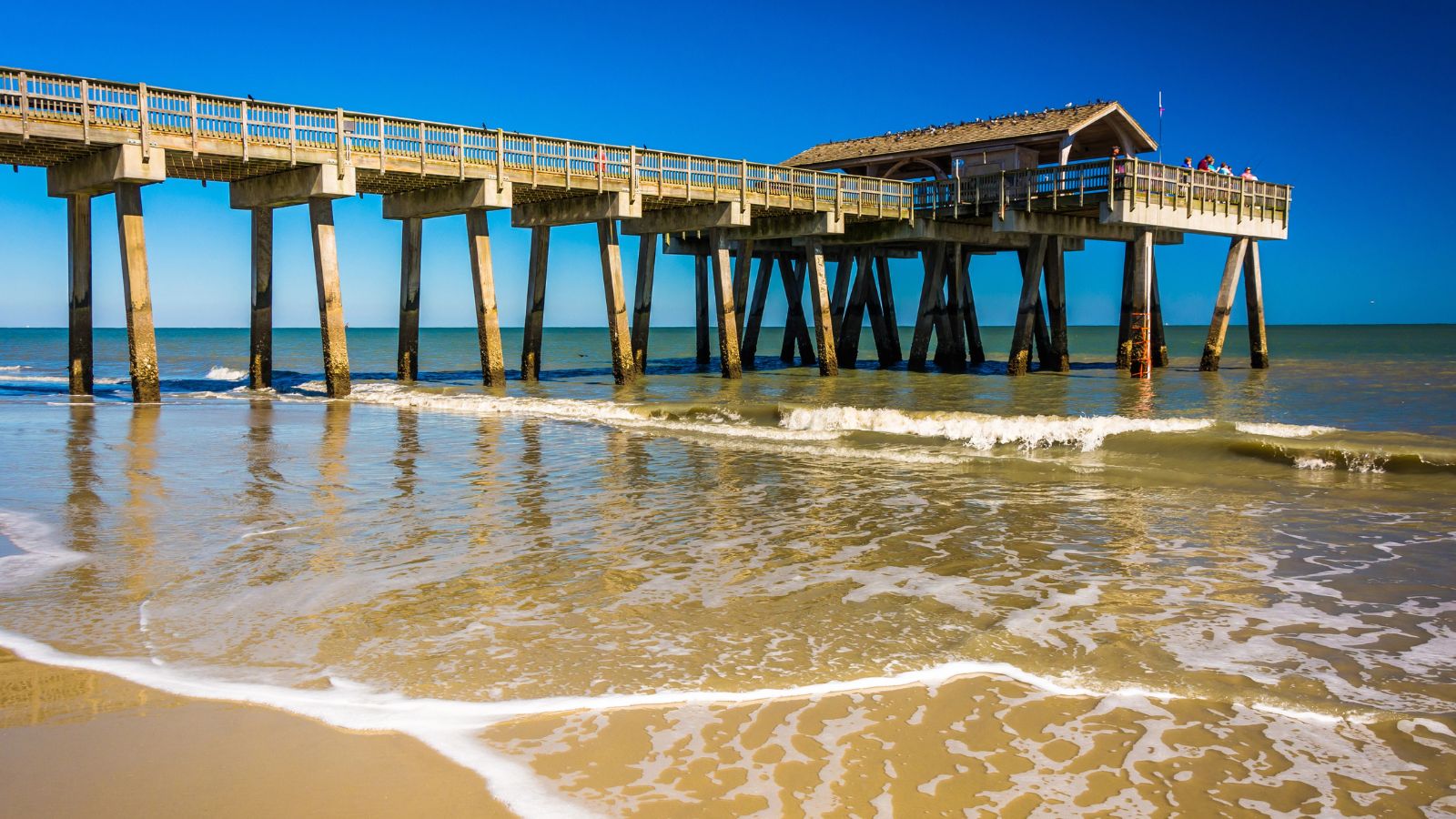 A wooden pier stretches over the ocean as waves meet a sandy beach beneath a clear blue sky.