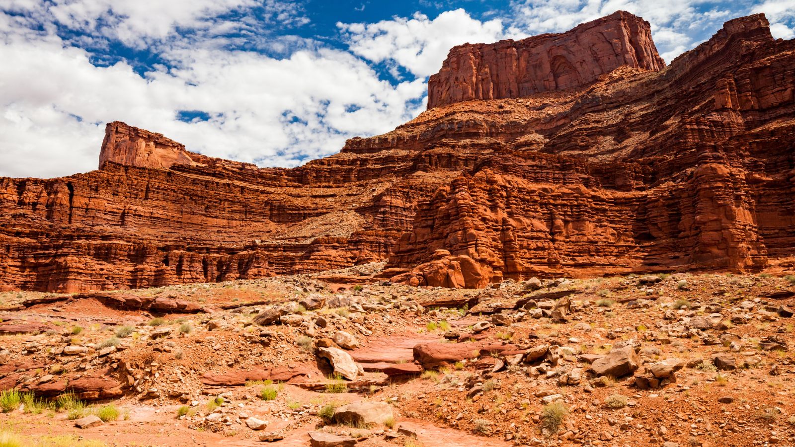 Red rock cliffs and mesas tower over a rocky, arid desert beneath a partly cloudy sky.