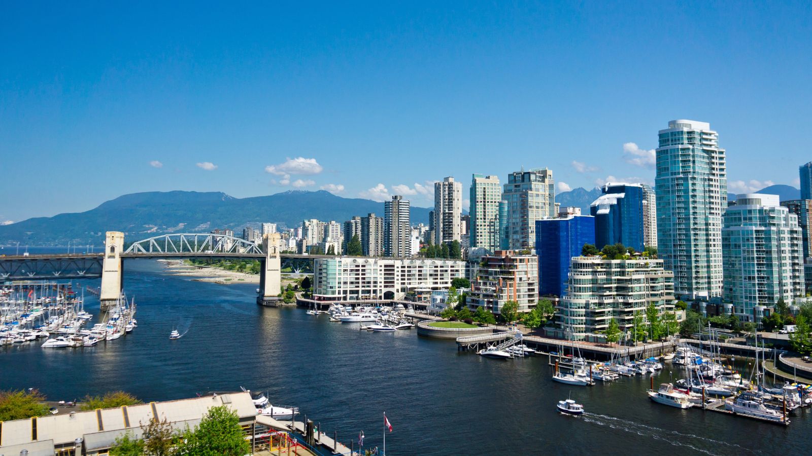 Vancouver waterfront with high-rises, marina boats, Burrard Street Bridge, and mountains under a clear blue sky.