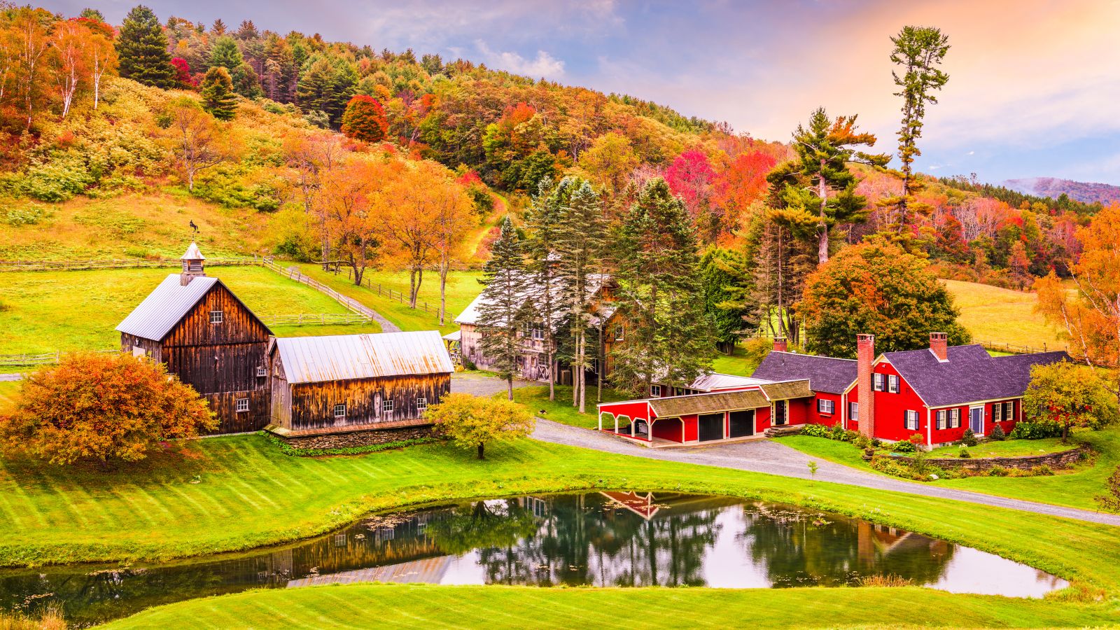 Farmhouse and barn beside a pond, surrounded by autumn trees on rolling hills under a partly cloudy sky.