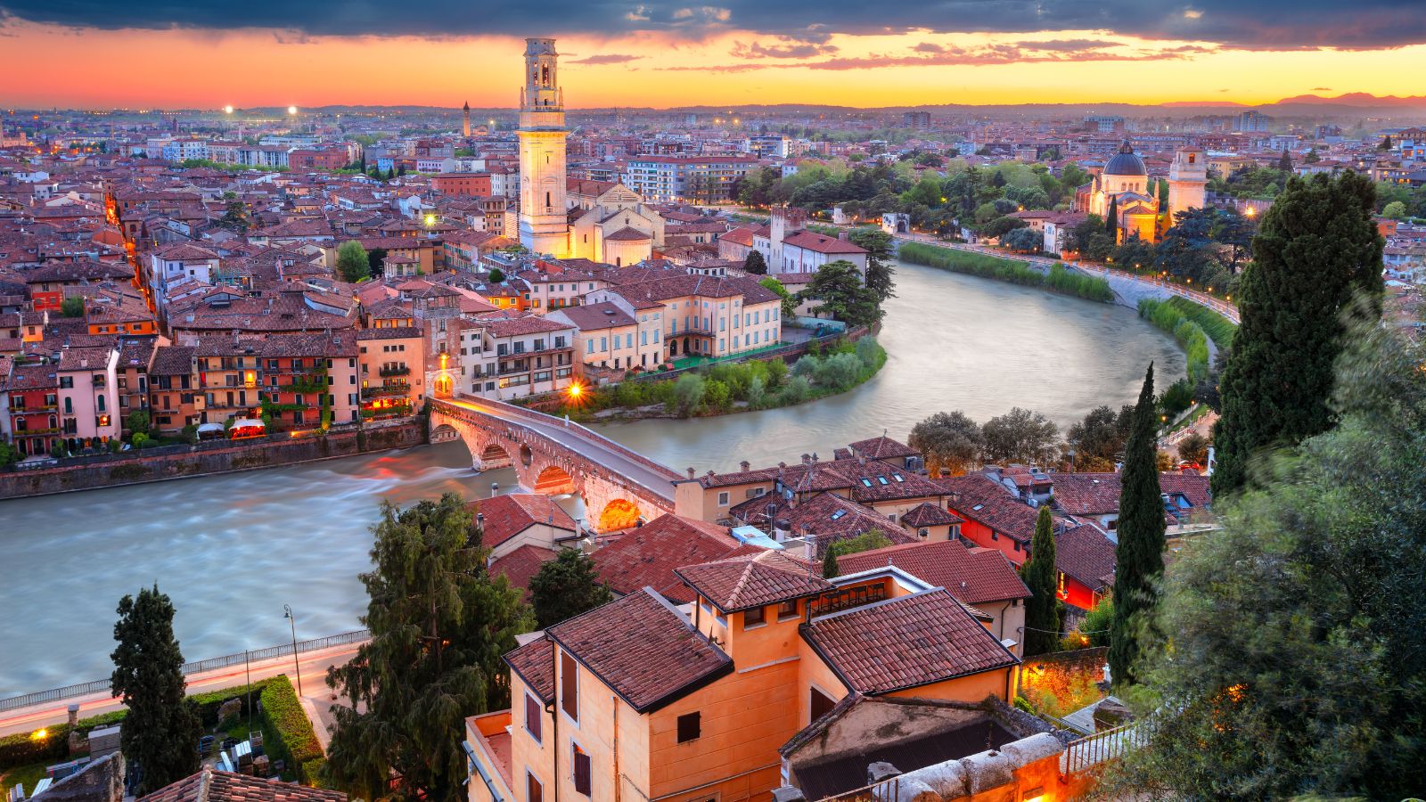 Verona at sunset with the Adige River winding past historic buildings and a stone bridge under a vibrant sky.