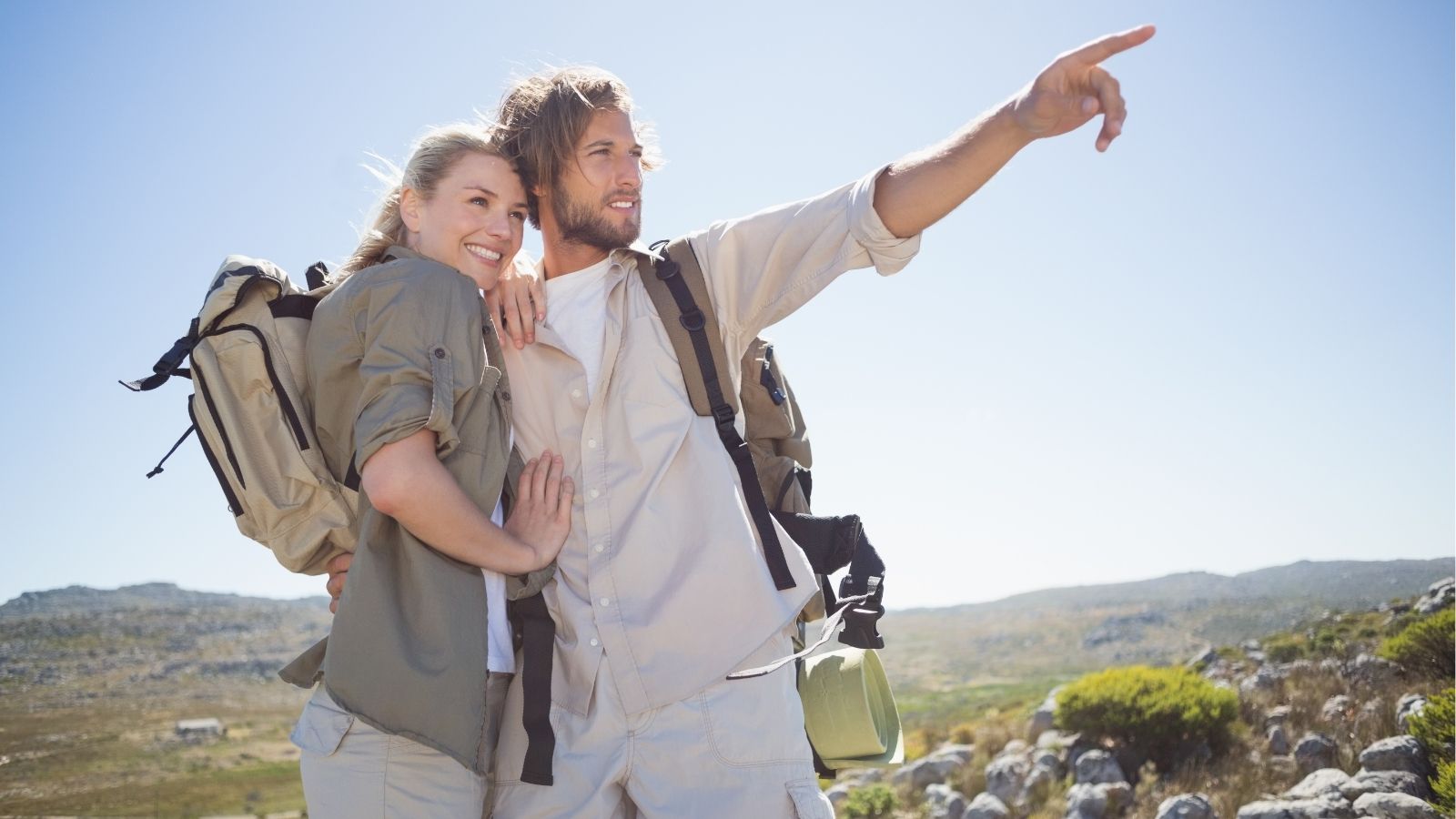 A man and woman in hiking gear stand on rocky terrain, the man pointing ahead as the woman smiles.