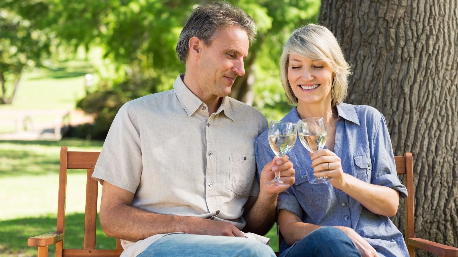A man and woman clink wine glasses while sitting on a wooden bench outside, surrounded by trees and greenery.