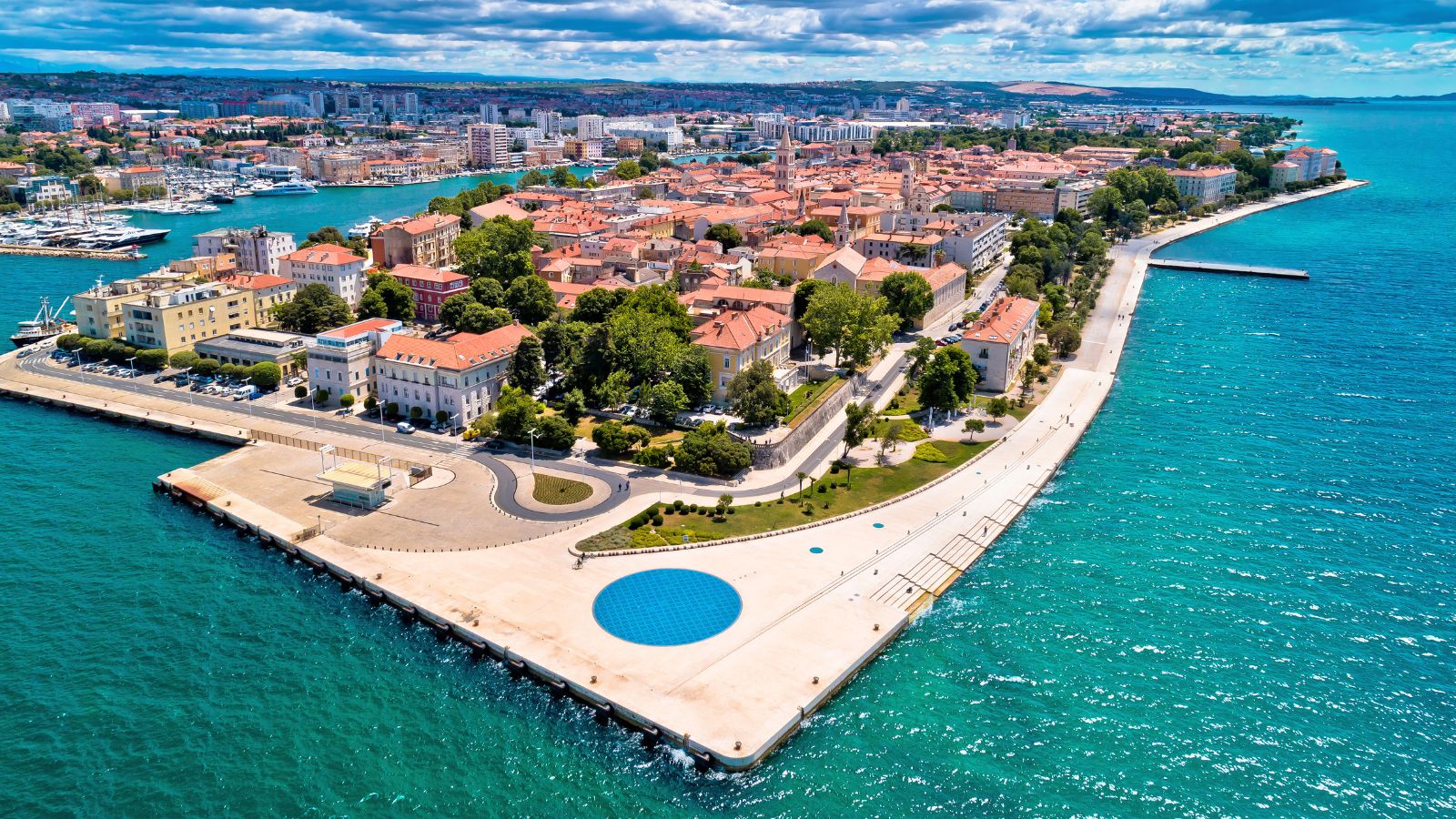 Aerial view of a coastal city with red roofs, a promenade, green trees, and turquoise water along the shore.