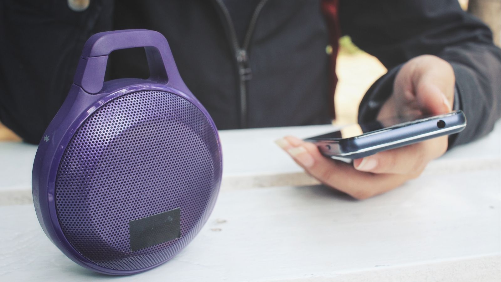 A person holds a smartphone beside a portable purple wireless speaker on a white surface.