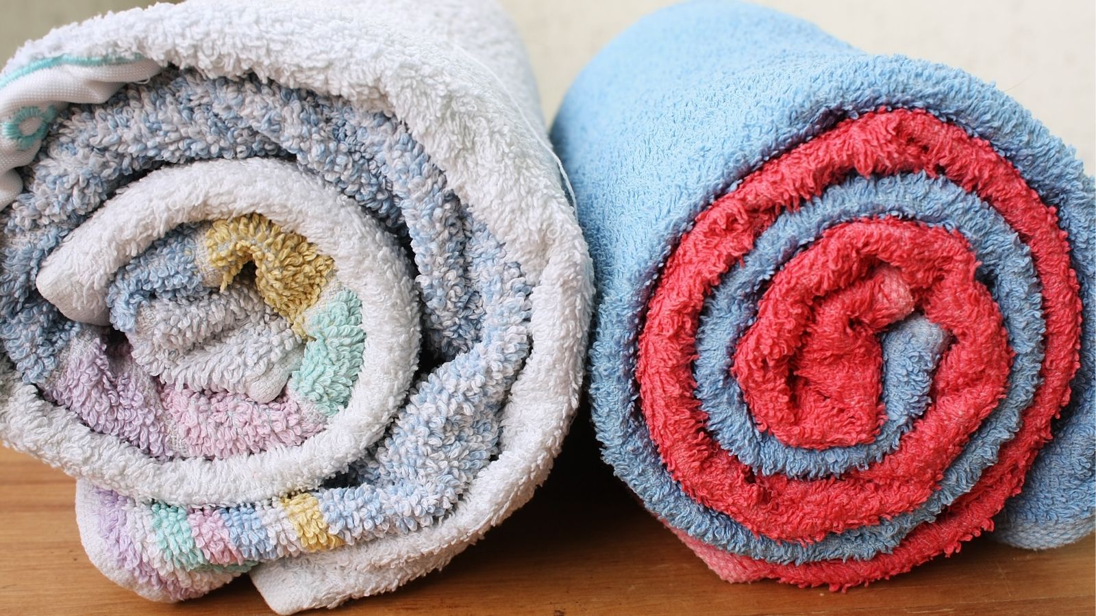 Two rolled bath towels sit side by side on wood; one is multicolored, the other is a blue towel rolled around a red towel.