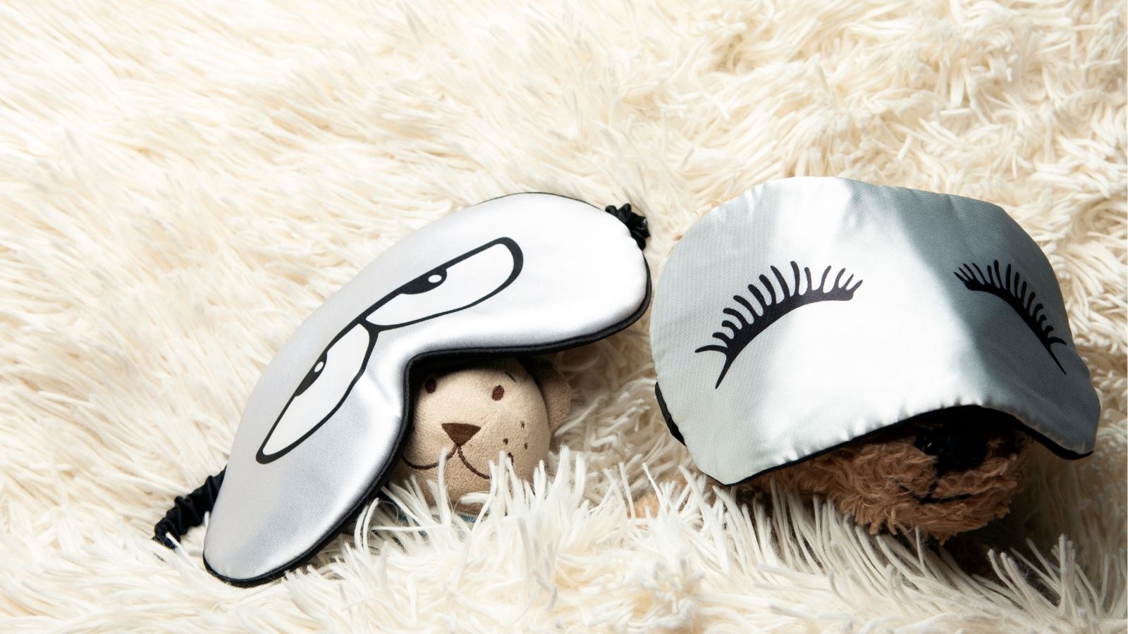 Two stuffed animals rest on a fluffy white surface, each in silver sleep masks—one with cartoon eyes, the other with eyelashes.