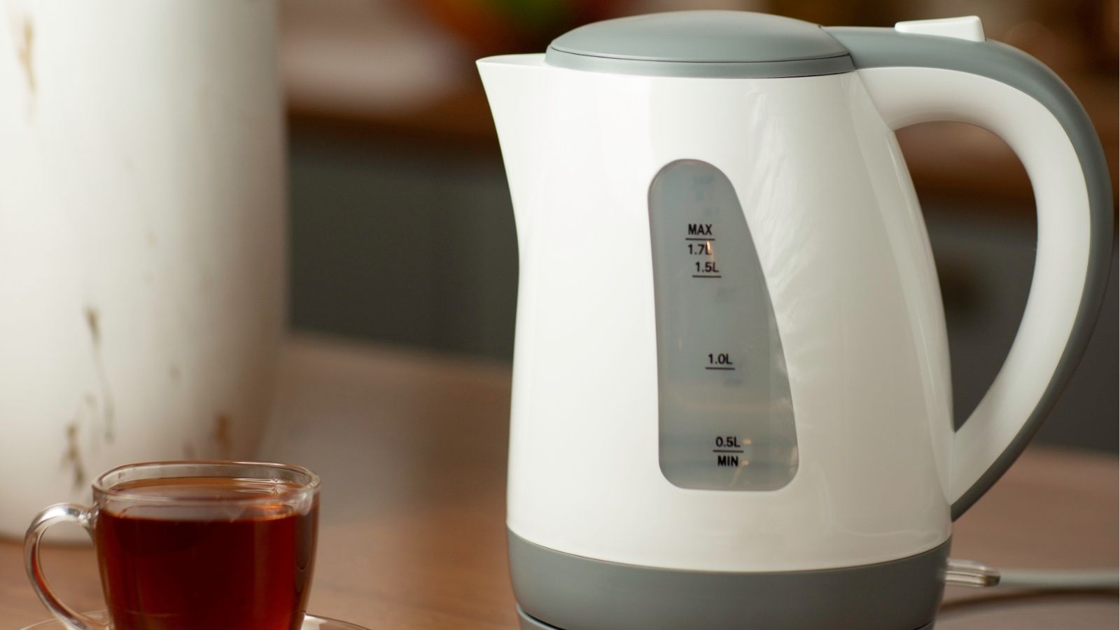 A white electric kettle with a water level indicator is on a counter beside a glass cup of tea.