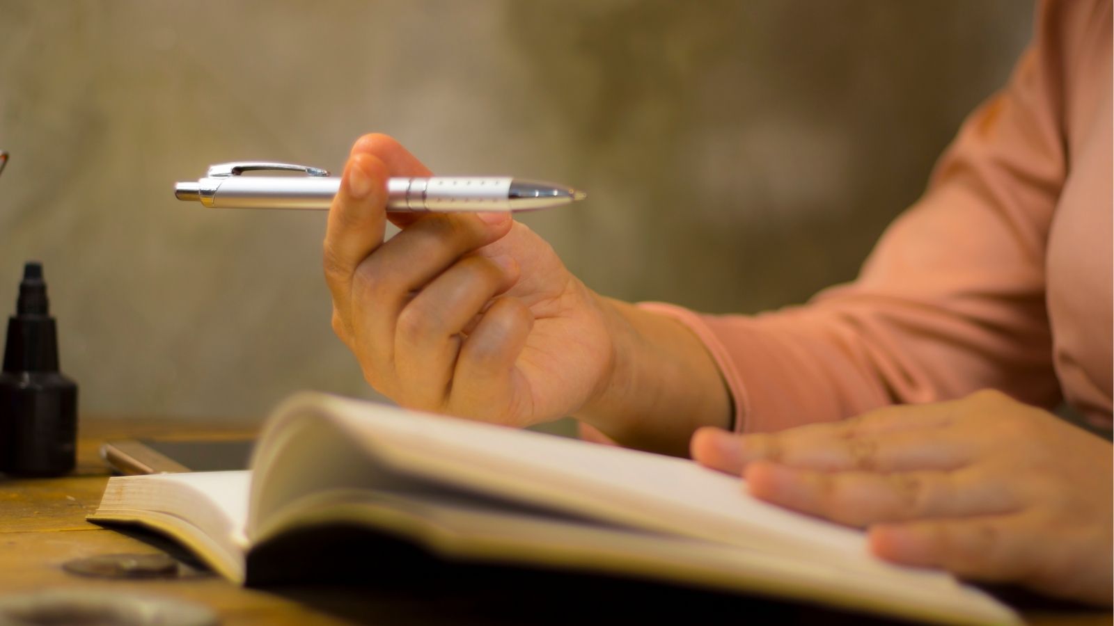 A person sits at a wooden desk, holding a pen and touching an open notebook.