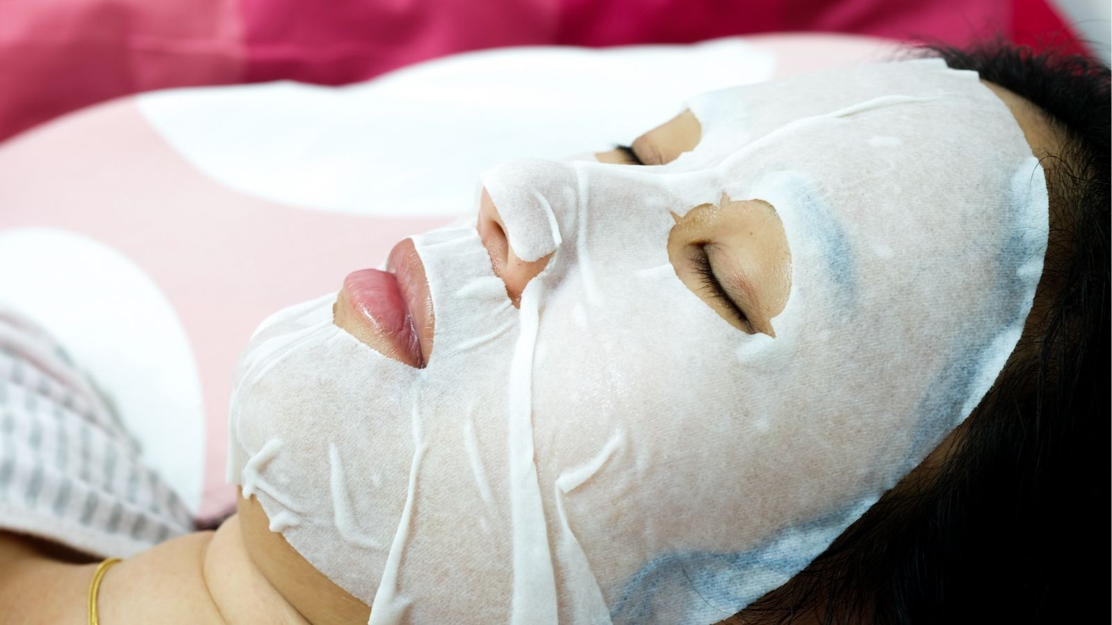 A person lies with eyes closed, wearing a white sheet face mask for skincare.