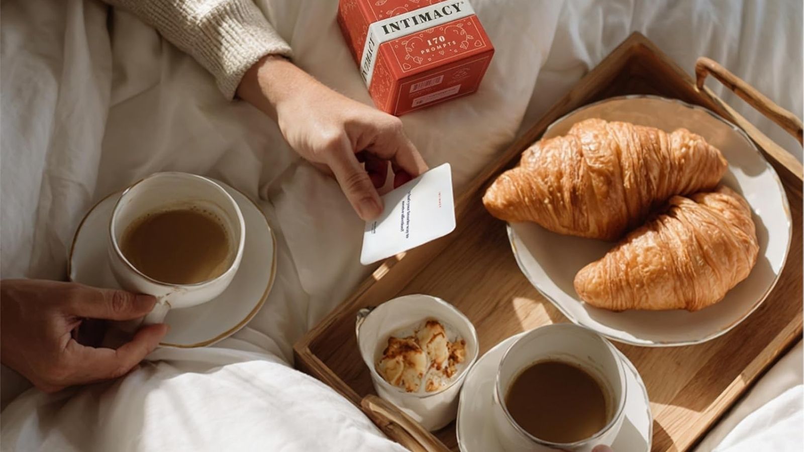 Two cups of coffee, croissants, and a small dish on a tray in bed; one person holds a packet marked “Intimacy.”.