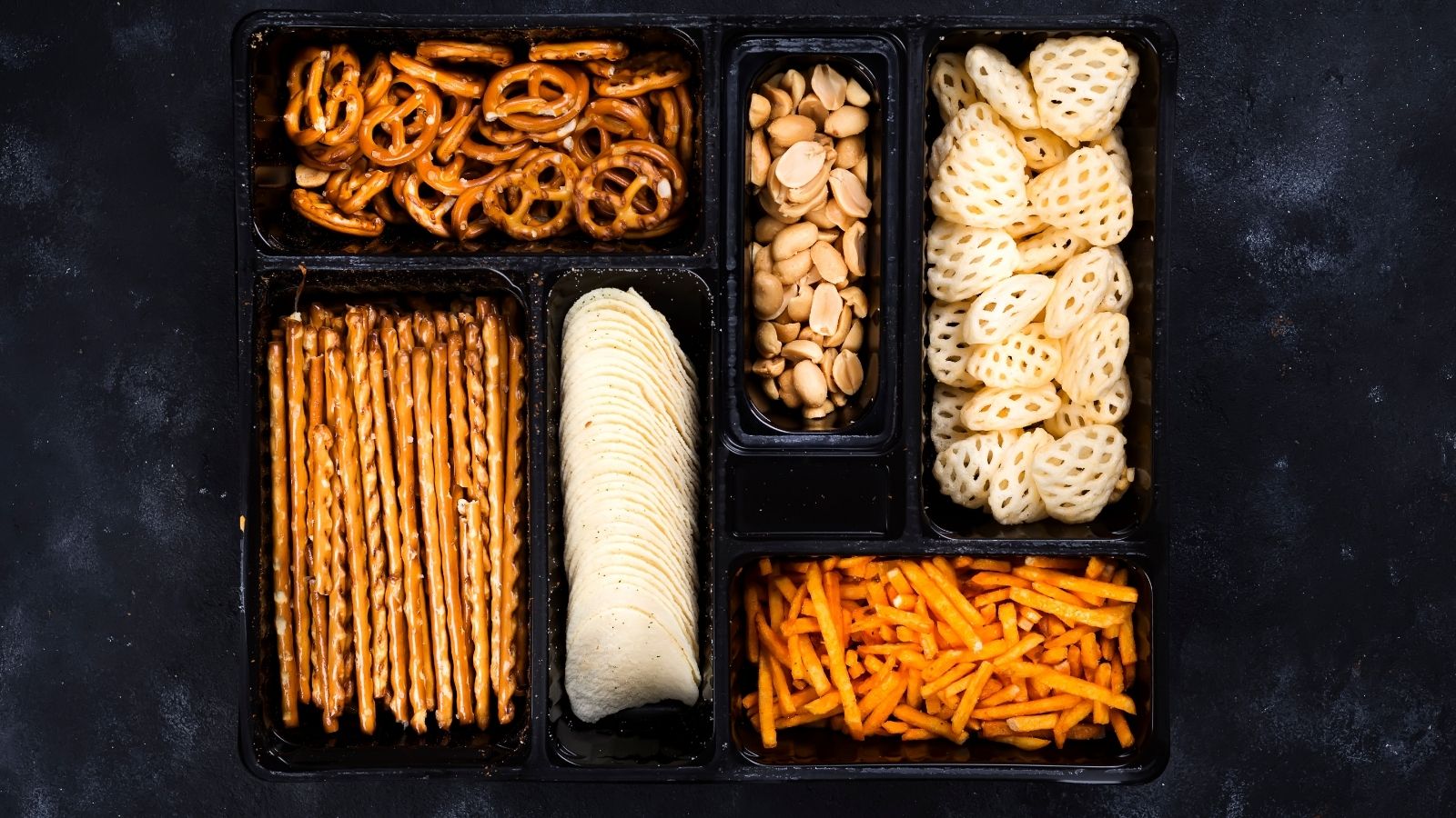 A black sectioned tray with pretzels, breadsticks, potato chips, cheese straws, lotus root chips, and peanuts on a dark surface.