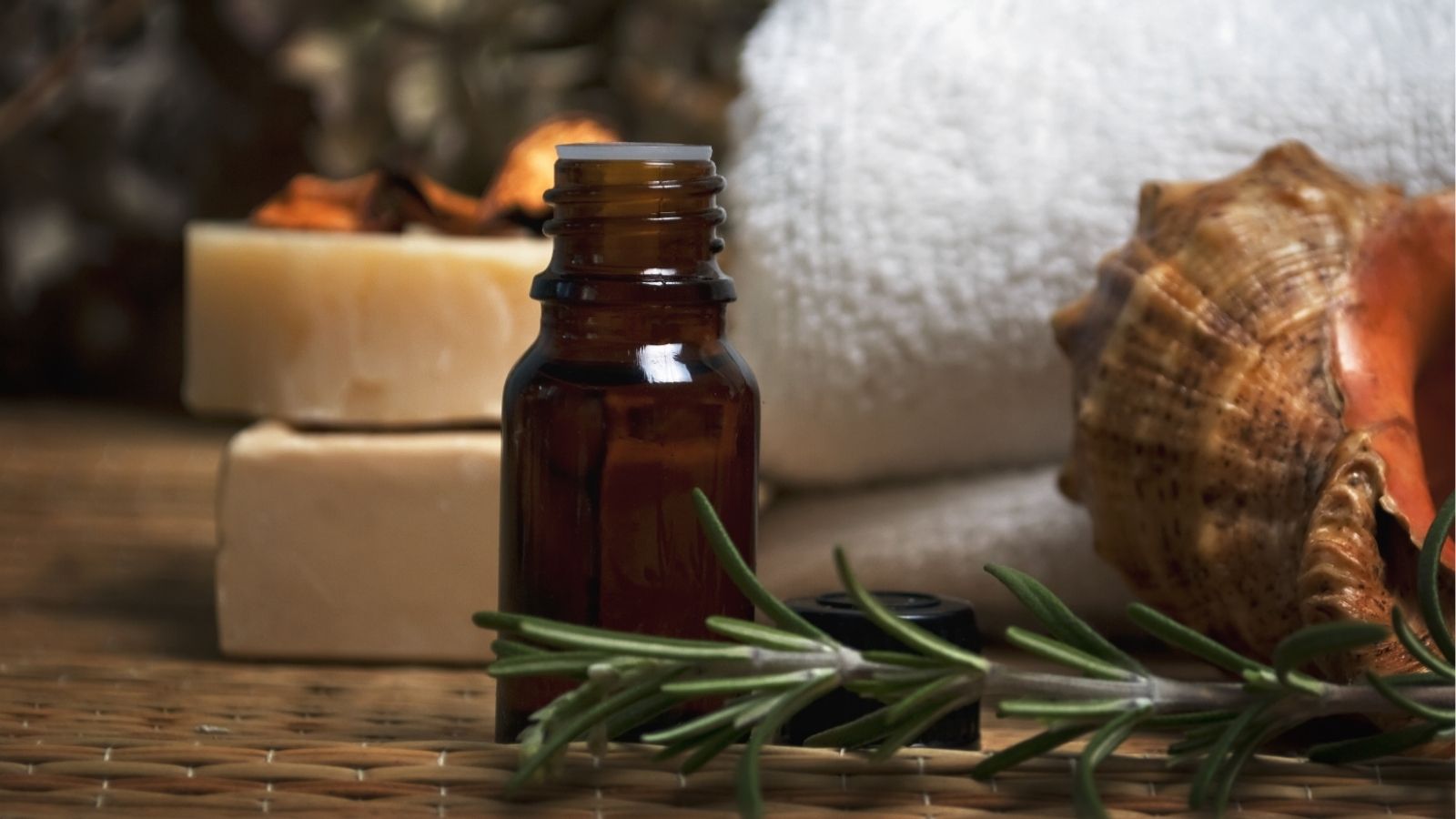 An amber glass bottle rests on a woven mat with soap bars, a white towel, a seashell, and rosemary nearby.
